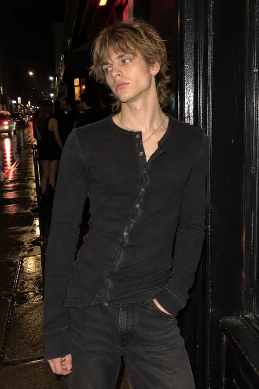 Concept render of ROOM 404: a man with messy medium hair leans against a dark doorway at night, wearing a washed-black deconstructed henley with an asymmetric placket and snap closures.